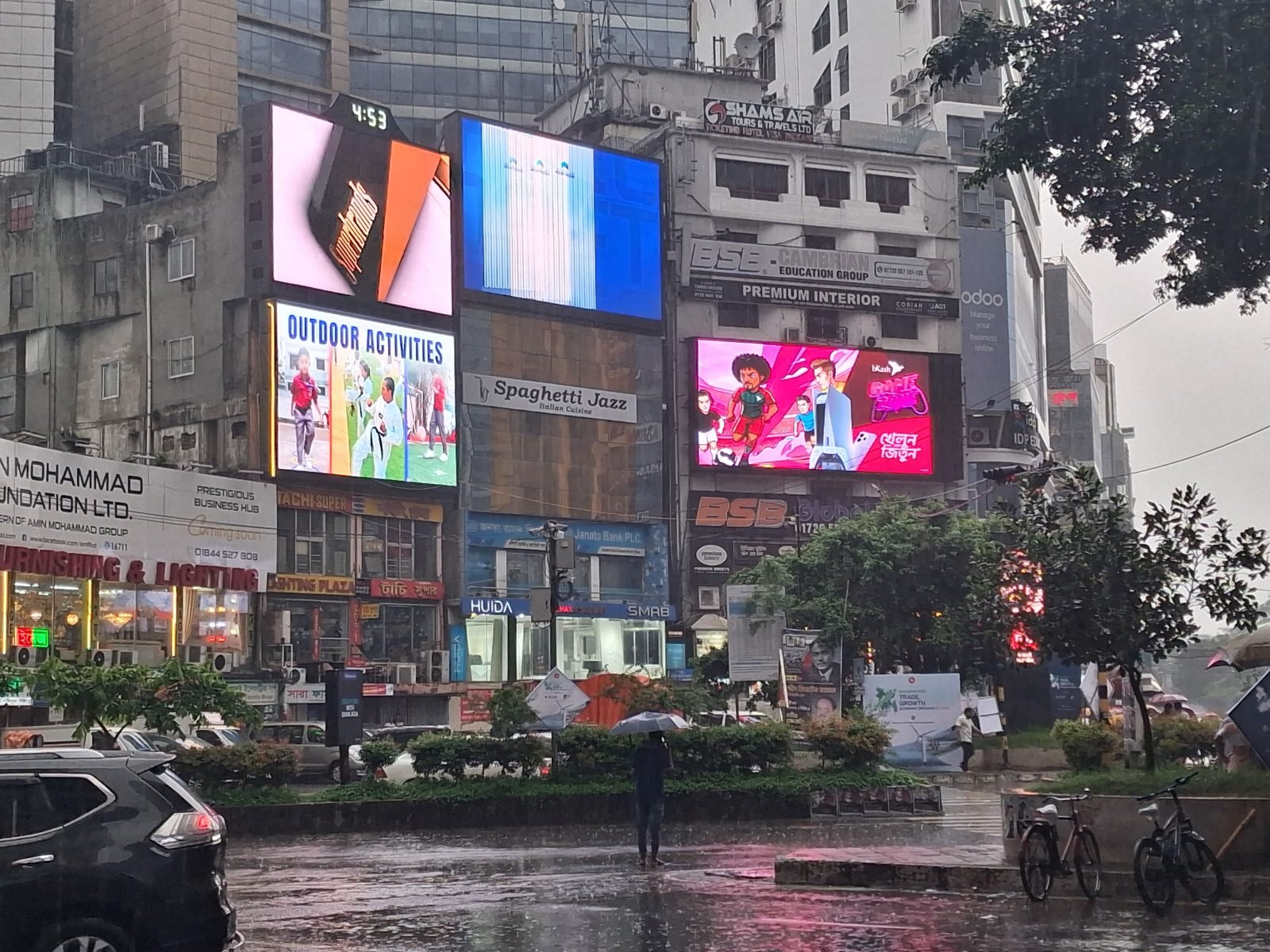 A live Bangla LED digital billboard cluster glowing over a busy Dhaka intersection at night